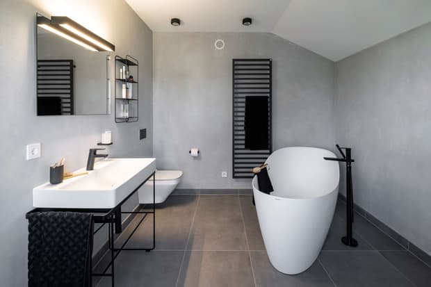 Modernes Badezimmer im Musterhaus mit freistehender Badewanne, grauen Fliesen, Design-Waschbecken und Wandheizkörper.