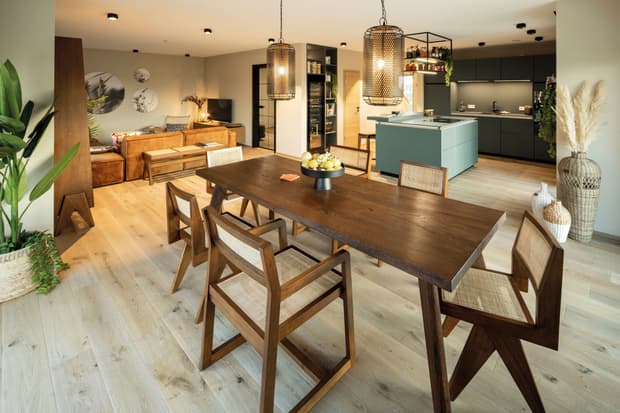 Offene Wohnküche im modernen Musterhaus mit Holztisch, Designerstühlen, Ledersofa, heller Beleuchtung und Kochinsel.