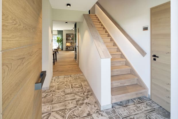 Heller Eingangsbereich im modernen Musterhaus mit Holztreppe, gemustertem Fliesenboden und offener Wohnraumansicht.