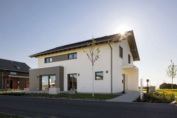 Modernes zweistöckiges Musterhaus mit Satteldach, Solarpanelen, heller Fassade und großen Fenstern bei Sonnenlicht.