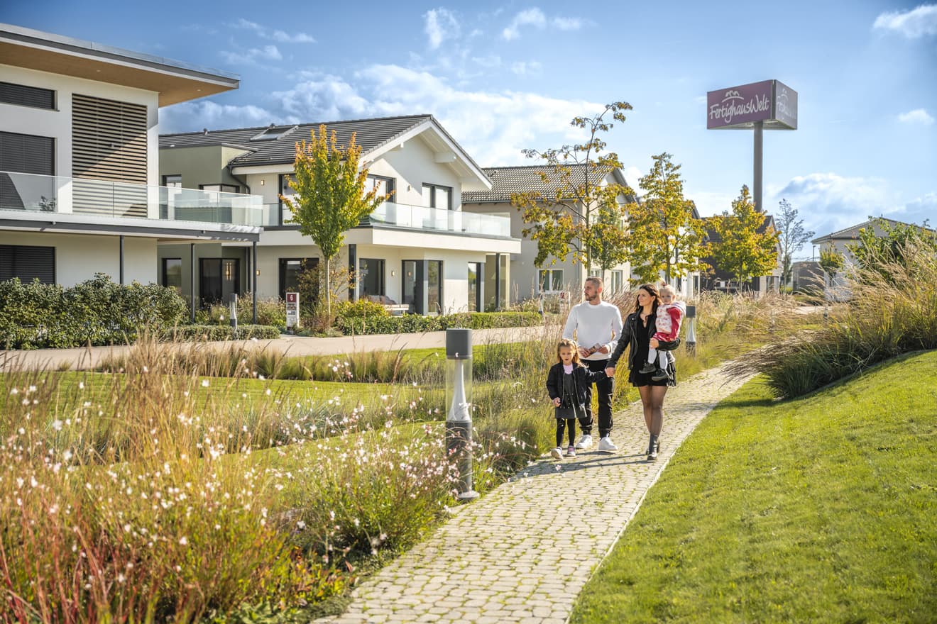Familie mit zwei Kindern spaziert bei Sonnenschein auf gepflastertem Weg durch moderne Fertighaussiedlung mit Grünflächen und Schild „FertighausWelt“.