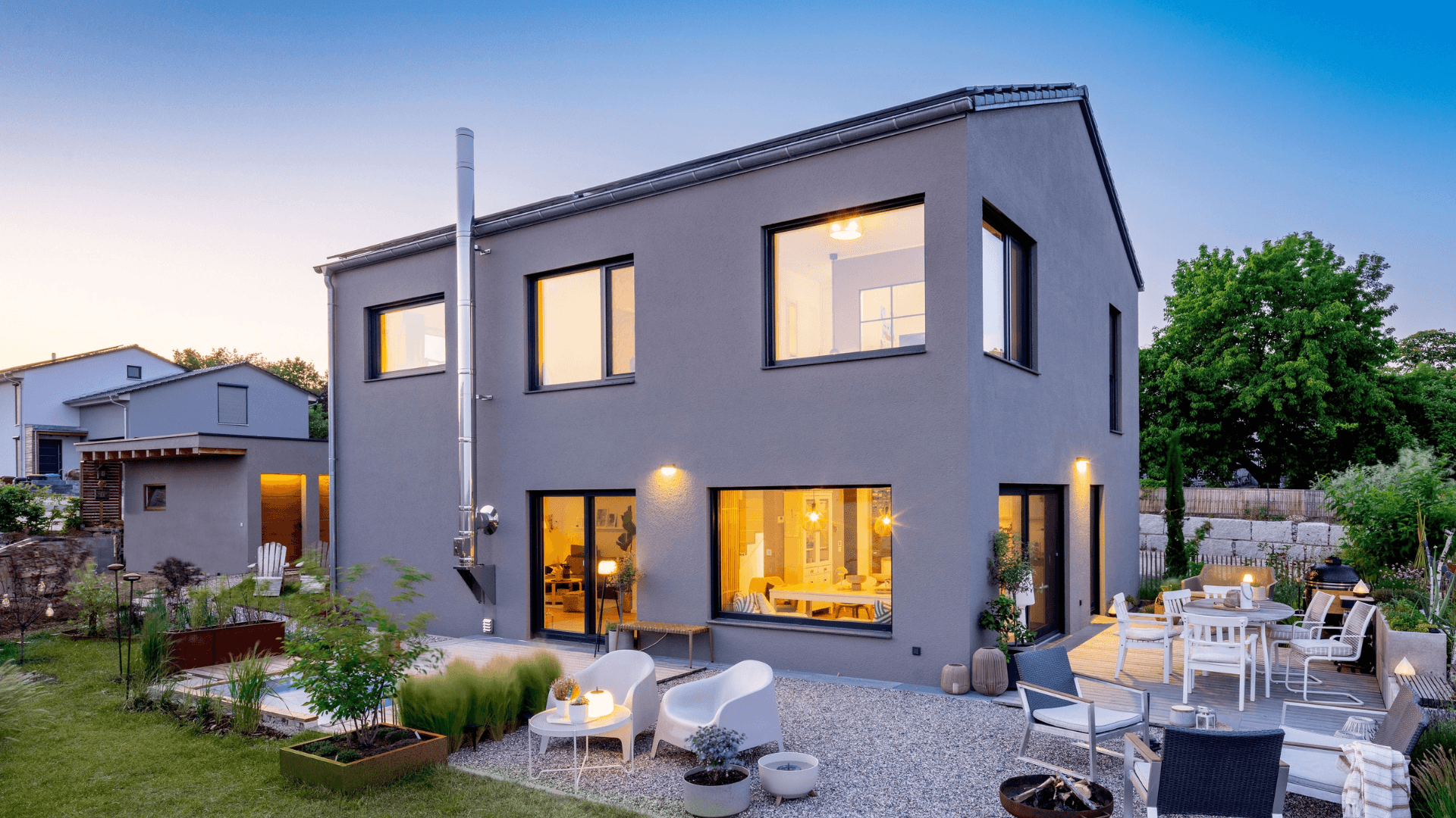 Modernes zweigeschossiges Einfamilienhaus mit grauer Fassade, großen Fenstern und beleuchteter Terrasse mit Gartenmöbeln und Rasenfläche bei Abenddämmerung