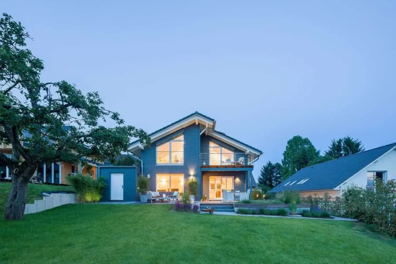 Modernes Musterhaus mit Satteldach, großer Fensterfront, beleuchteter Terrasse und gepflegtem Garten in ruhiger Wohnlage.