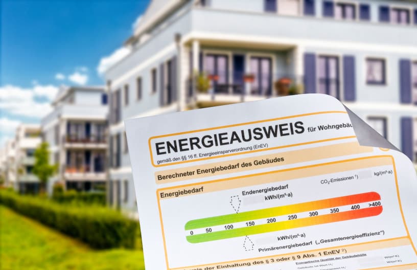 Energieausweis für Wohngebäude vor modernem Mehrfamilienhaus, zeigt Energieeffizienzskala und berechneten Energiebedarf.