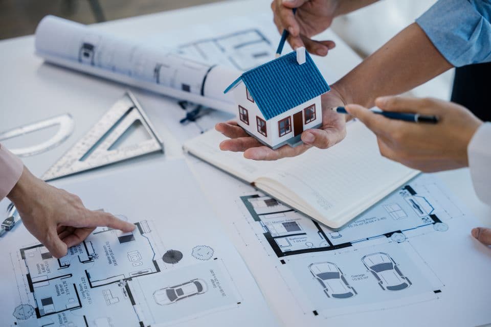 Architekten planen Musterhaus mit Grundrissen, Notizbuch und Miniaturhausmodell auf Tisch im modernen Planungsbüro.