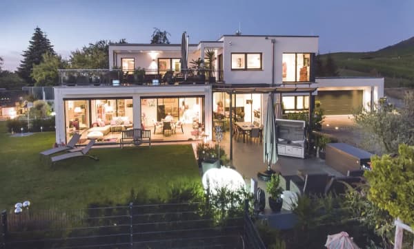 Luxus-Fertighaus mit Flachdach, großer Glasfront, Terrasse, Balkon, Garten und stilvoller Beleuchtung bei Dämmerung.