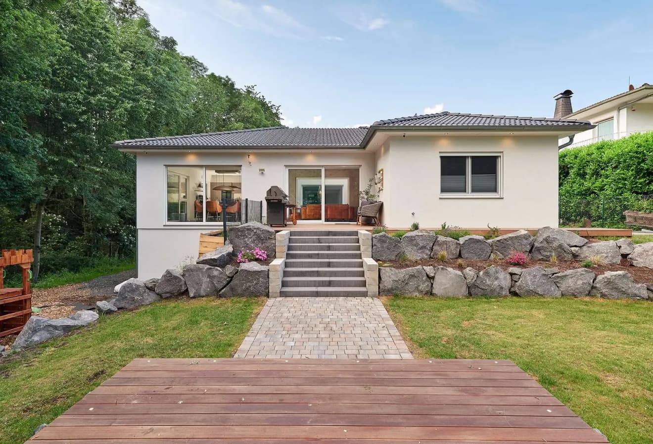 Luxhaus Bungalow Walmdach 172: Barrierefreier Winkelbungalow mit Natursteinmauer, Holzterrasse und gepflegtem Garten. Modernes Einfamilienhaus in Hanglage – komfortables Wohnen auf einer Ebene.
