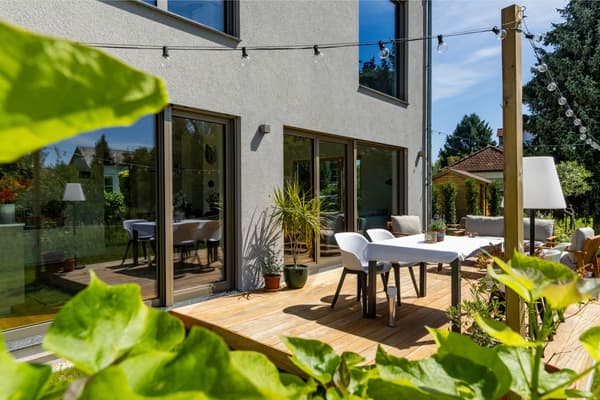 LUXHAUS Terrasse mit Holzdeck, Esstisch und Lichterkette vor bodentiefen Fenstern, umgeben von grünem Garten