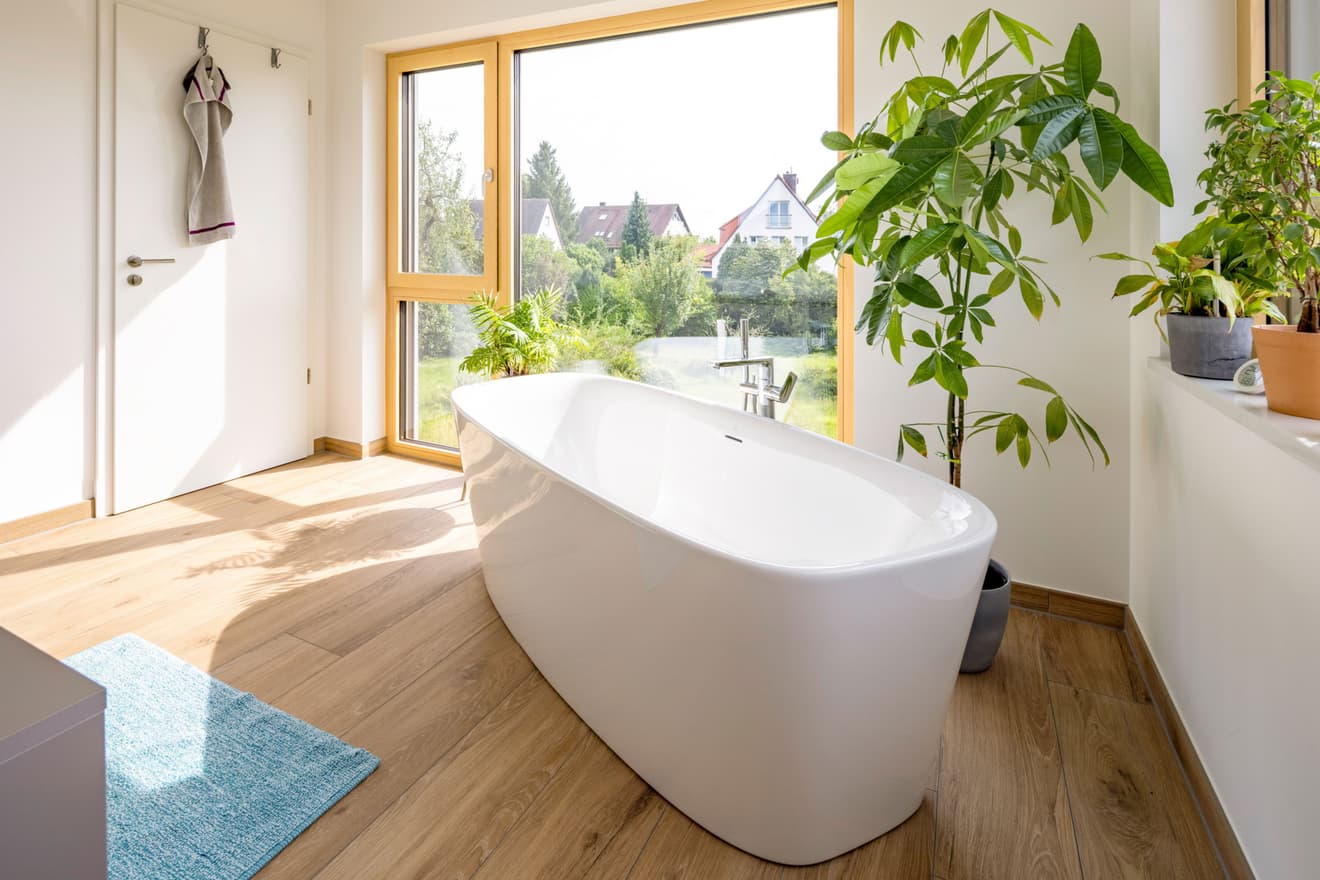 Modernes Badezimmer im LUXHAUS mit freistehender Badewanne, großem Panoramafenster und Holzboden in heller Atmosphäre