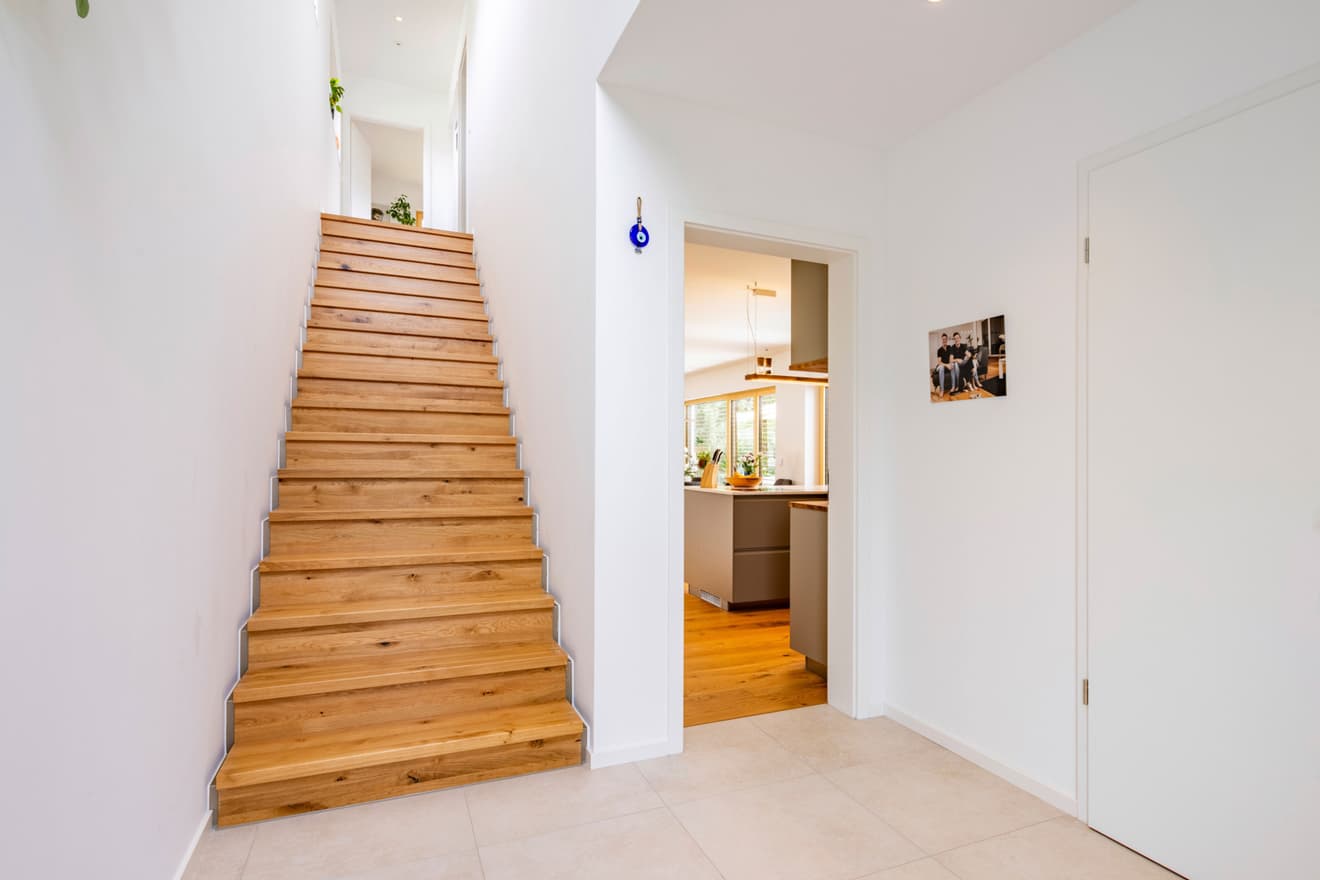 Moderner Flur im LUXHAUS mit Holztreppe aus Eiche, weißen Wänden und Durchblick zur offenen Küche im Hanghaus