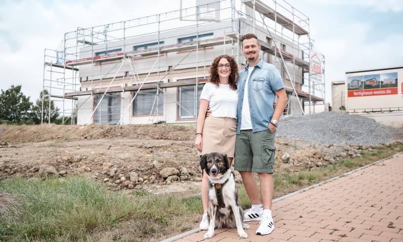 Junges Paar mit Hund vor einem im Bau befindlichen Einfamilienhaus mit Gerüst und Baustellenumgebung