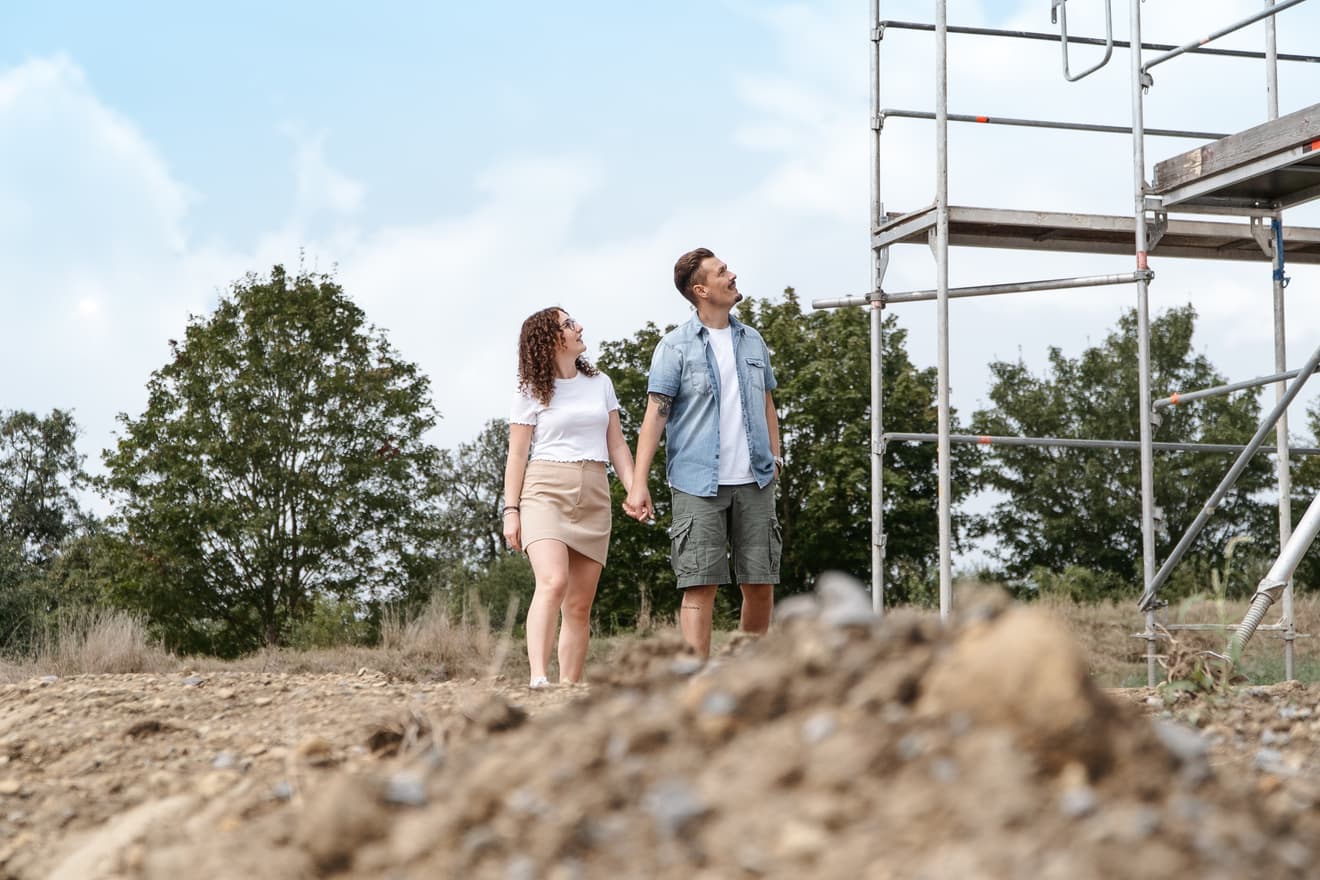 Paar hält Händchen auf Baustelle und blickt zum Gerüst bei bewölktem Himmel und Bäumen im Hintergrund