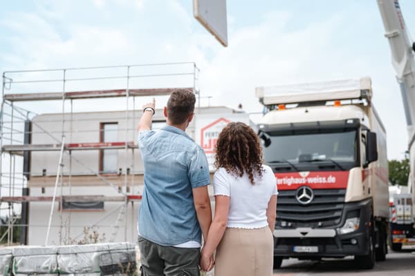 Junges Paar hält sich an der Hand und betrachtet Rohbau eines Fertighauses mit LKW und Gerüst im Hintergrund