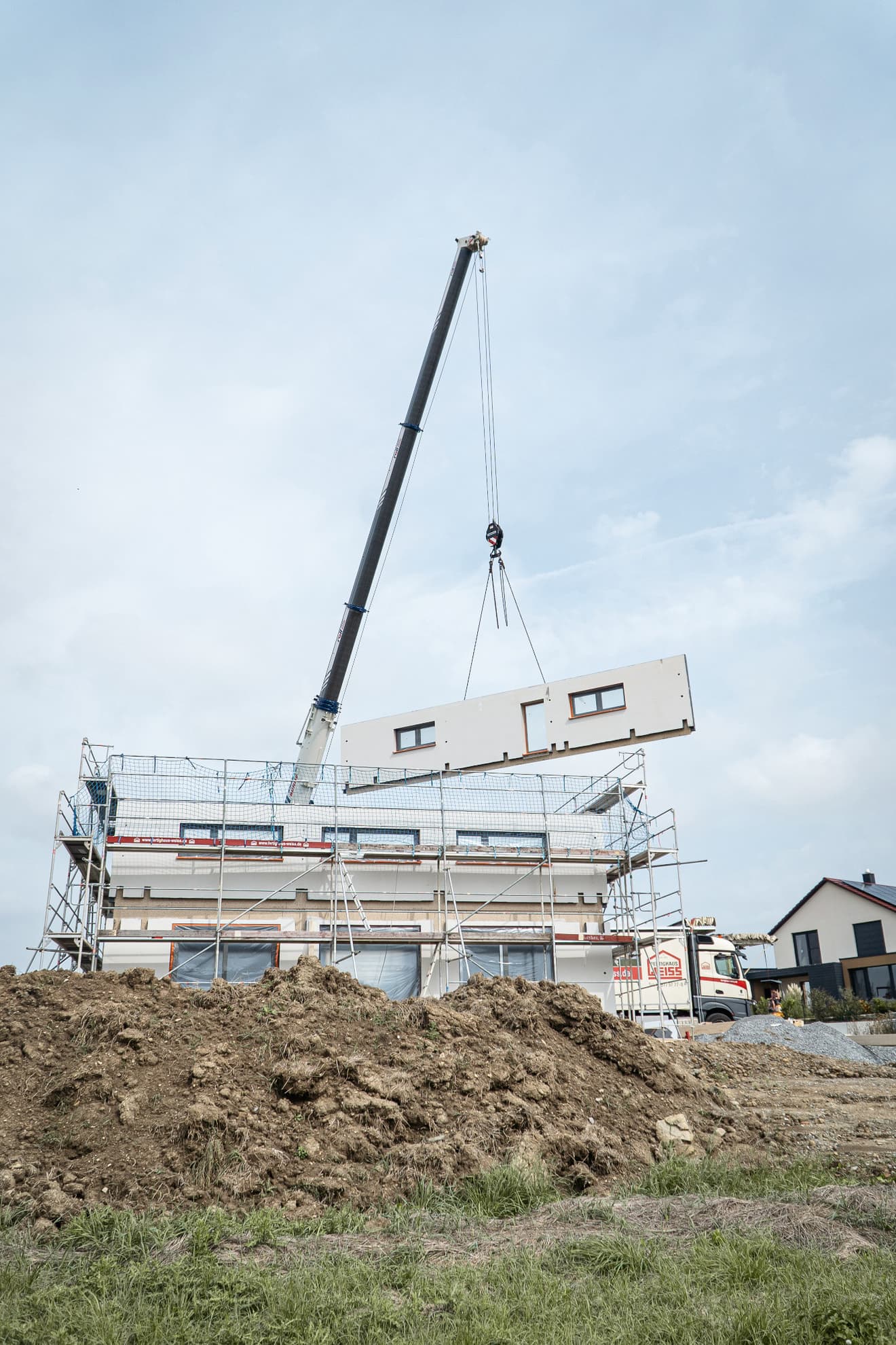 Baukran hebt weiße Fertigbauelement mit Fenster und Tür auf ein eingerüstetes Haus auf Baustelle