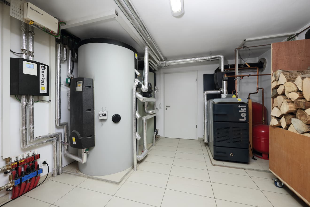 Moderner Heizungsraum mit Warmwasserspeicher, Pelletofen, Lüftungsrohren und gestapeltem Brennholz