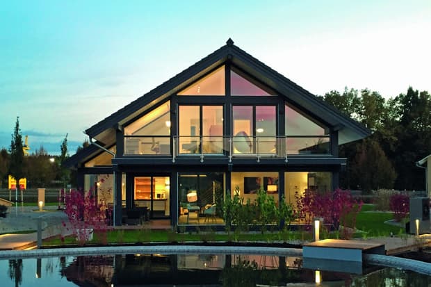Modernes Haus mit großflächigen Glasfenstern und Beleuchtung bei Dämmerung mit Teich und Garten im Vordergrund