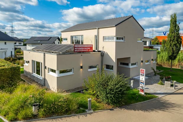 Modernes Musterhaus mit Flachdach und Solaranlage, beige Fassade, Gartenanlage und Hinweisschild zur Neueröffnung.