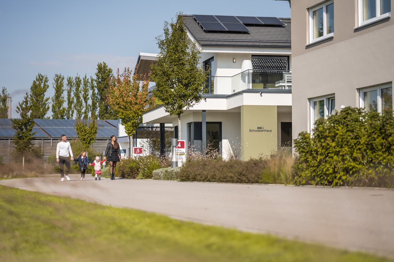 Familie mit zwei Kindern spaziert vor modernem weißen Haus mit Solarpaneelen und Glasbalkon an sonnigem Tag