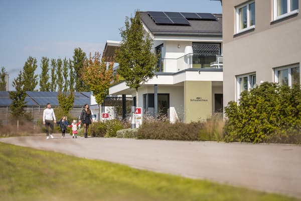 Familie mit zwei Kindern spaziert vor modernem weißen Haus mit Solarpaneelen und Glasbalkon an sonnigem Tag