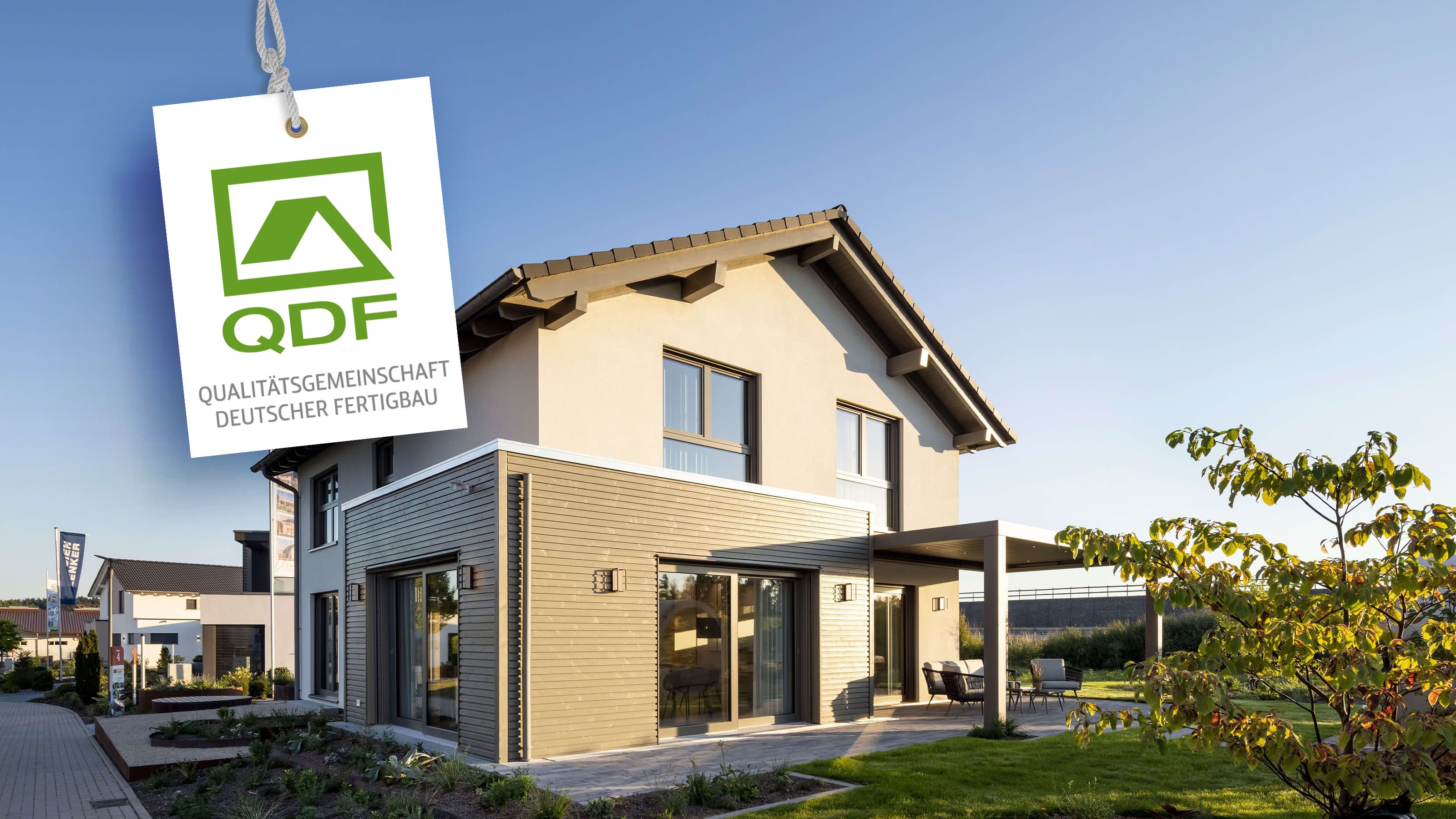 Modernes Einfamilienhaus mit Terrasse und Garten im Sonnenlicht; links hängt ein QDF‑Qualitätssiegel.