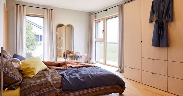 Helles Schlafzimmer im modernen Musterhaus mit großem Bett, Holzschrank, Spiegel, Vorhängen und Blick ins Grüne.