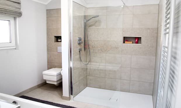 Modernes Badezimmer im Musterhaus mit begehbarer Dusche, Glaswand, Wandnischen, hellen Fliesen und hängendem WC.