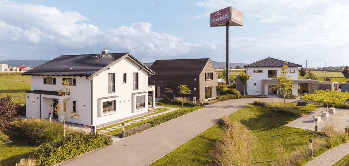 Luftaufnahme moderner Musterhäuser mit gepflegten Gärten, Straße und hohem Werbeschild im Hintergrund.