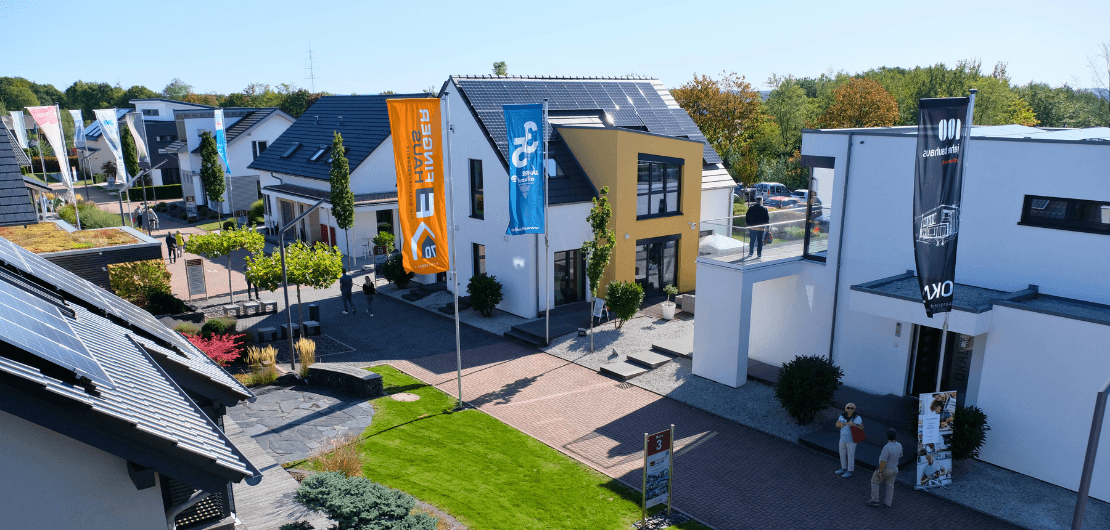 Blick auf moderne Musterhäuser mit Solarpanels, Fahnen, Wegen und Besuchern zwischen den Gebäuden.
