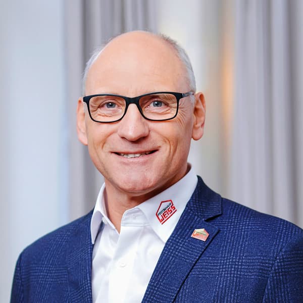 Porträt eines älteren Mannes mit Brille, weißem Hemd und blauem Sakko vor hellgrauem Hintergrund