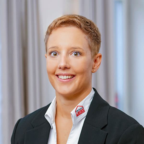 Porträt einer lächelnden Frau mit kurzen blonden Haaren, weißem Hemd und schwarzem Blazer vor hellem Hintergrund