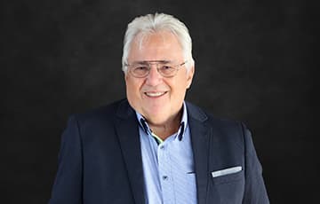 Älterer Mann mit weißen Haaren und Brille trägt dunkelblauen Anzug und hellblaues Hemd vor schwarzem Hintergrund
