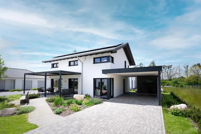 Modernes zweistöckiges Musterhaus mit Carport, Terrasse, Flachdach, Gartenanlage und heller Fassade unter blauem Himmel.