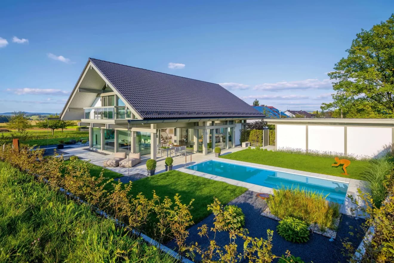 Modernes Luxus-Fertighaus mit Satteldach, großer Glasfront, Pool, Terrasse, Garten und Blick auf die umliegende Landschaft.
