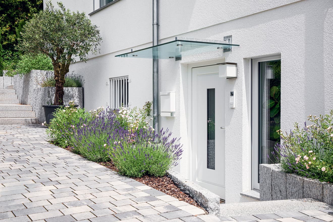 Moderne weiße Haustür mit Glasvordach und gepflegtem Vorgarten. Zeitgenössische Hauseingangsgestaltung mit Lavendel, Pflastersteinen und minimalistischem Design für ansprechende Außenarchitektur.