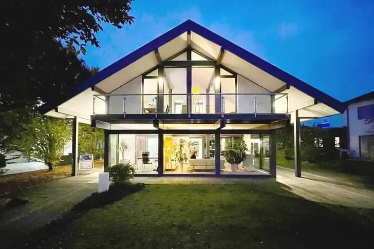 Modernes Musterhaus mit großer Glasfassade, Satteldach und eleganter Abendbeleuchtung – stilvolle Architektur mit hellem Wohngefühl.