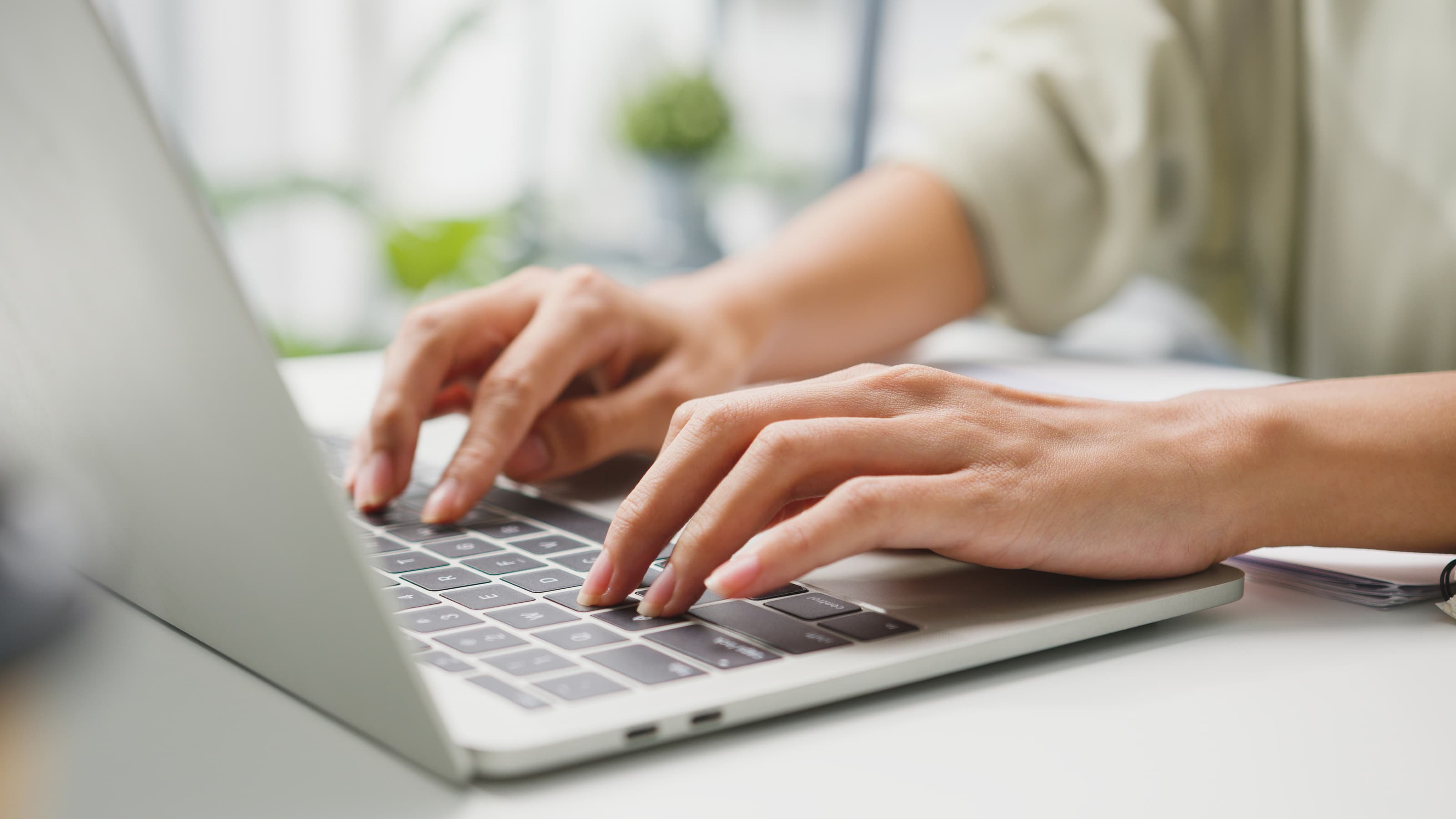 Person tippt mit beiden Händen auf silbernem Laptop in hellem Arbeitsumfeld mit Pflanzen im Hintergrund
