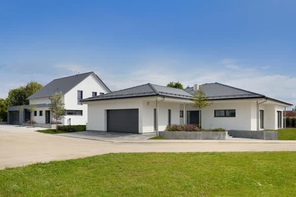 Modernes weißes Einfamilienhaus mit dunklem Walmdach und Garage, davor Einfahrt und Rasen.
