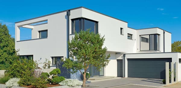 Modernes zweigeschossiges Flachdachhaus in kubischer Architektur mit weißer Putzfassade und großen, dunklen Fensterflächen. Rechts befindet sich eine Doppelgarage mit grauen Toren, links ein überdachter Terrassenbereich mit offener Betonrahmung.