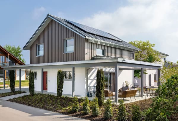 Modernes zweigeschossiges Einfamilienhaus mit Satteldach und Solarpaneelen; Obergeschoss mit grauer Holzfassade, Erdgeschoss weiß verputzt. Überdachte Terrasse mit Sitzgruppe und Esstisch.