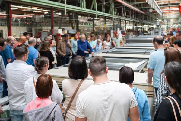 Gruppe von Besuchern steht in einer großen Produktionshalle und hört einem Mitarbeiter zu, der einen Fertigungsprozess erklärt