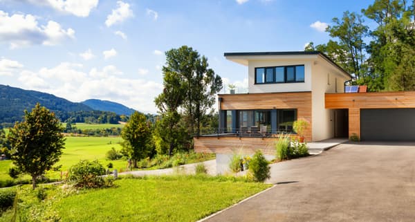 Modernes zweigeschossiges Einfamilienhaus mit Flachdach, weißer Putzfassade und Holzverkleidung im Obergeschoss; große Fensterfronten, Terrasse mit Sitzmöbeln und angrenzender Doppelgarage