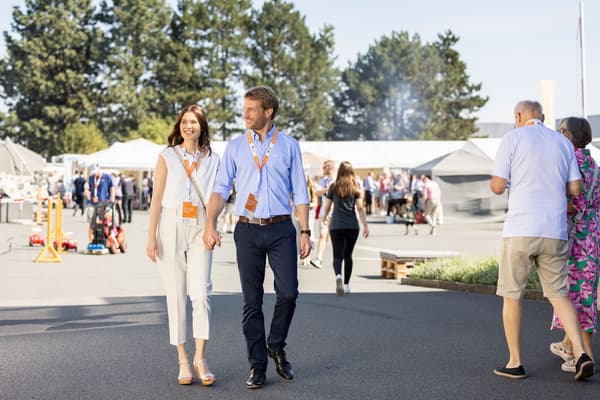 Ein Paar mit orangefarbenen Besucher-Ausweisen geht lächelnd Hand in Hand über ein sonniges Veranstaltungsgelände mit vielen Menschen, weißen Pavillons und Infoständen im Hintergrund.