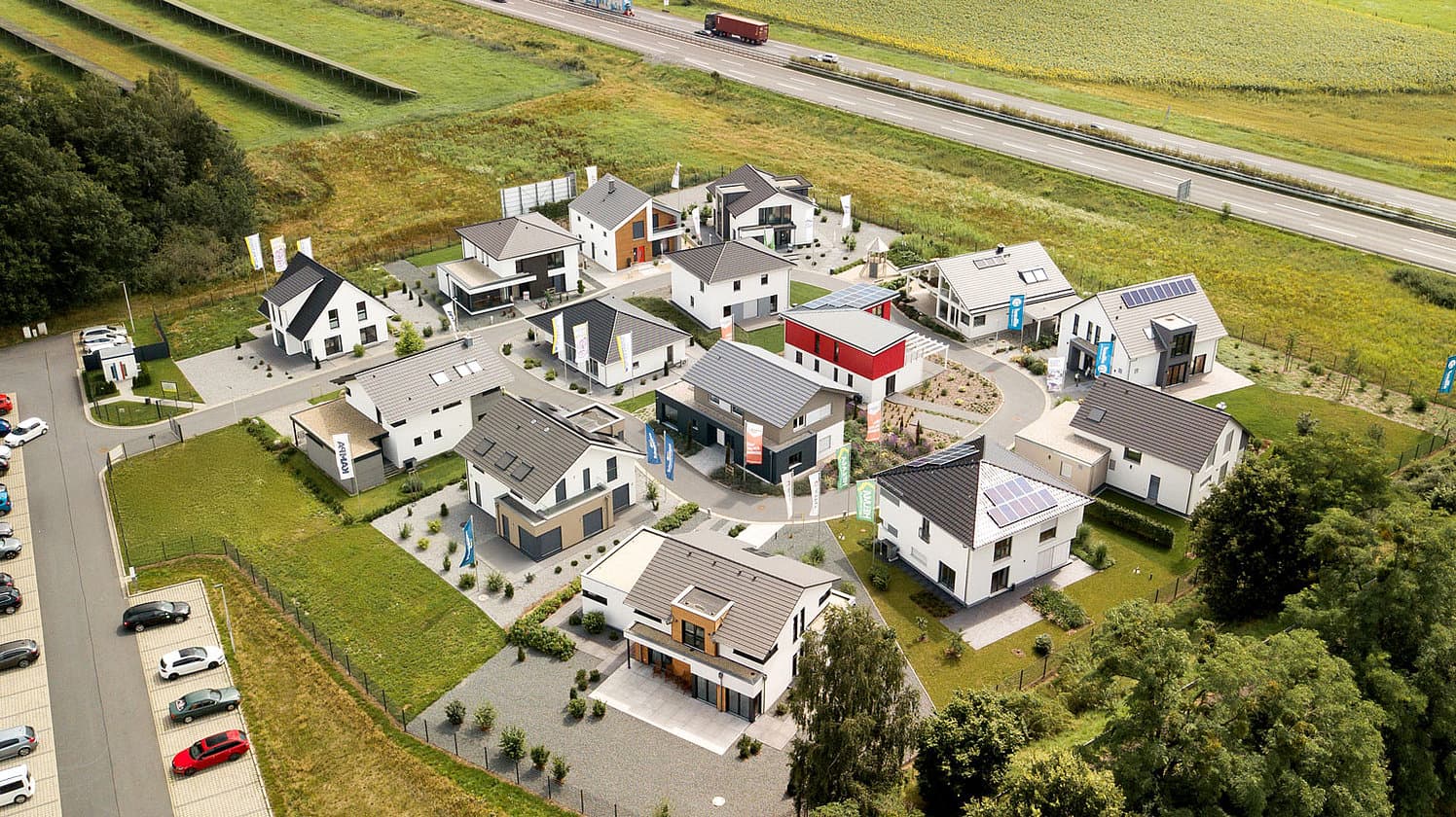 Luftaufnahme einer modernen Musterhaussiedlung mit Einfamilienhäusern, Solarzellen, grünem Umland und Autobahn im Hintergrund