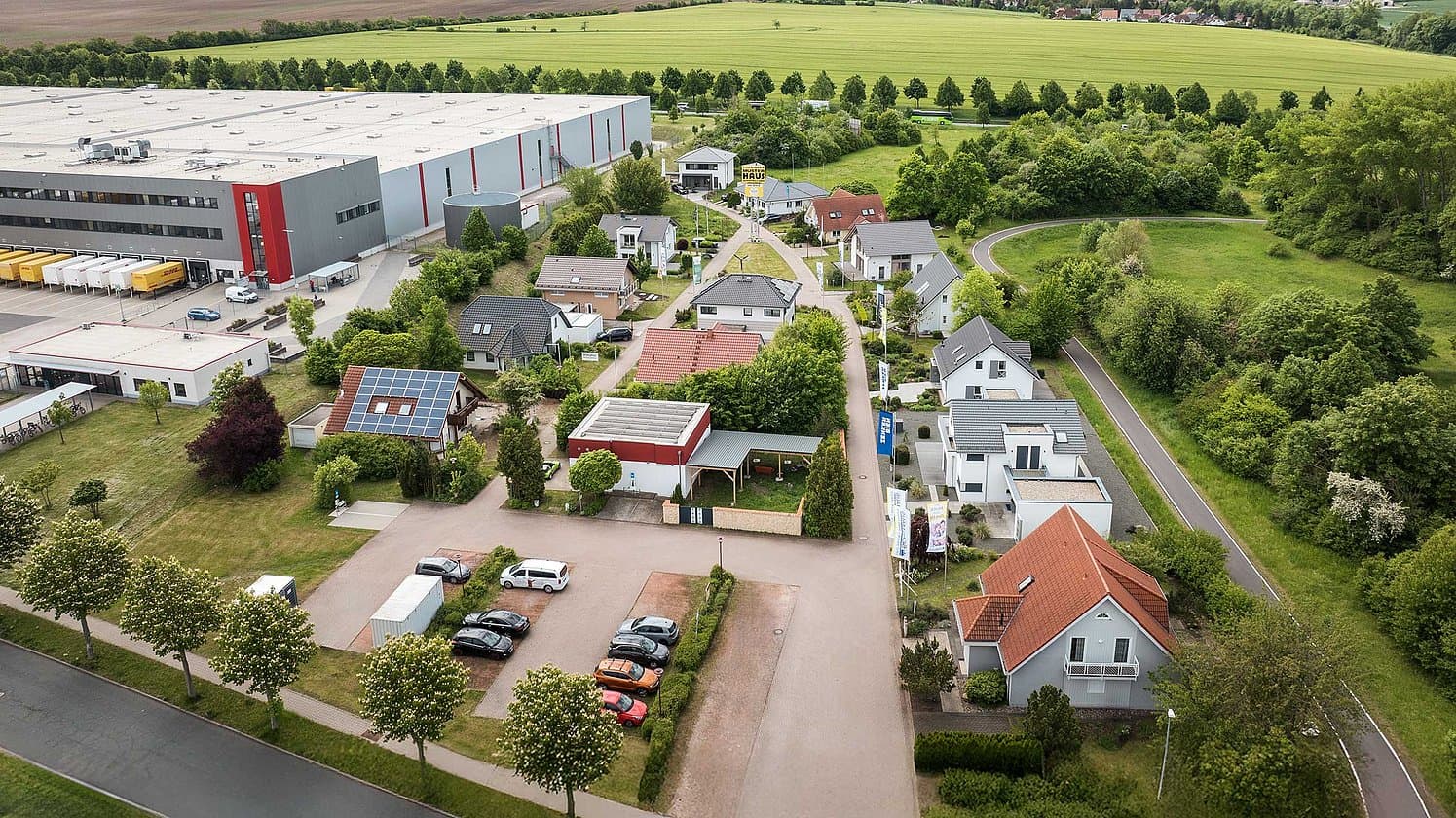 Luftaufnahme eines modernen Wohngebiets mit Einfamilienhäusern, Solaranlagen und angrenzendem Gewerbegebiet in ländlicher Umgebung