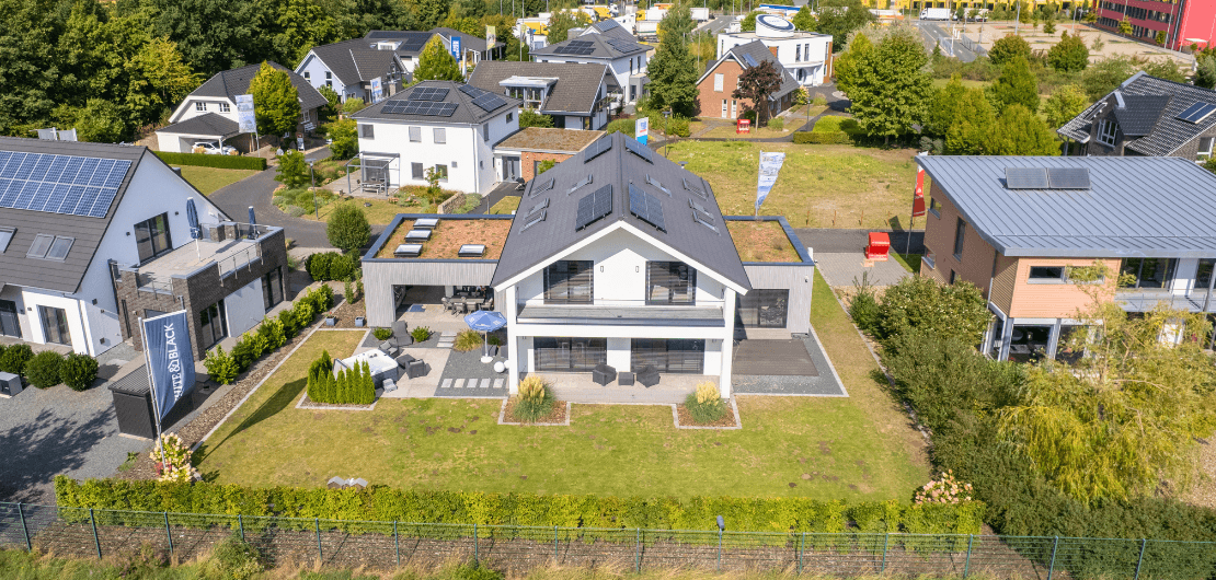 Luftaufnahme eines modernen Hauses mit Solarpanels, Terrasse, Garten und Nachbarhäusern im Grünen.